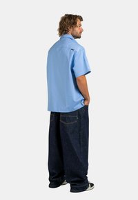 Man with curly hair wearing a light blue short-sleeve shirt and loose dark blue jeans, standing with hands in pockets, facing sideways.