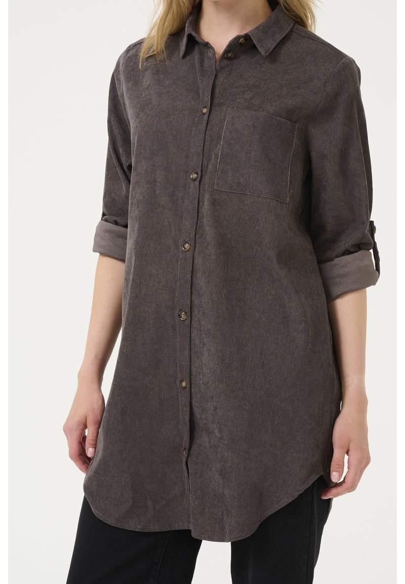 Chemise gris foncé à manches longues avec boutons à l’avant, poche poitrine et manches retroussées portée par une personne devant un fond uni.