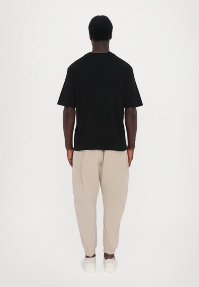 Schwarzes kurzärmliges T-Shirt mit lockerer Passform, kombiniert mit beigen schmal zulaufenden Hosen und weißen Sneakers, von hinten betrachtet.