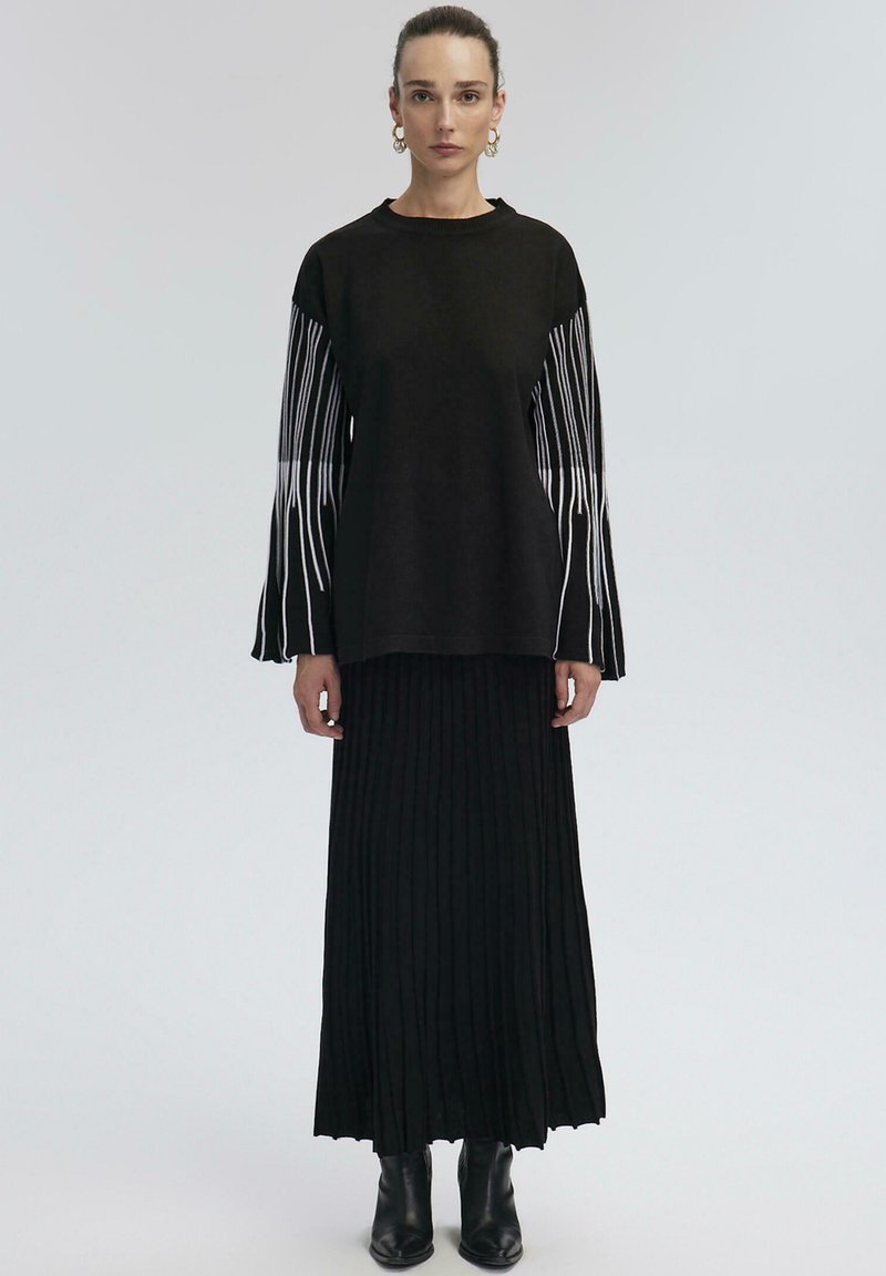 Black knit top with white vertical stripes on sleeves, paired with a long black pleated skirt and black ankle boots. Simple, modern design.