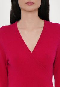 Pulover înfășurat fucsia cu mâneci lungi, realizat din țesătură tricotată moale. Prezintă un decolteu adânc în V și detalii subtile texturate de-a lungul cusăturilor.