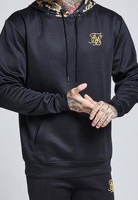 Sudadera con capucha negra con logo dorado, textura suave y capucha con diseño en el interior. Cuenta con un bolsillo delantero y cordones.