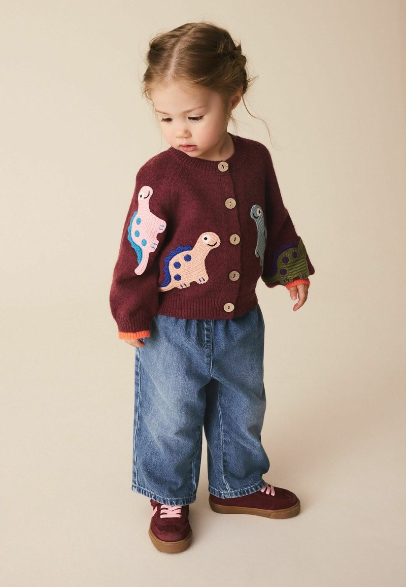 Niño pequeño con cárdigan granate con parches de dinosaurios coloridos, vaqueros azules holgados y zapatos granate con cordones rosas, mirando hacia abajo.