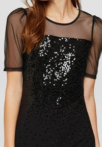 Černé šaty s průhlednými síťovanými krátkými rukávy, zdobené flitry vytvářejícími gradientní efekt. Měkká textura a přiléhavá silueta.