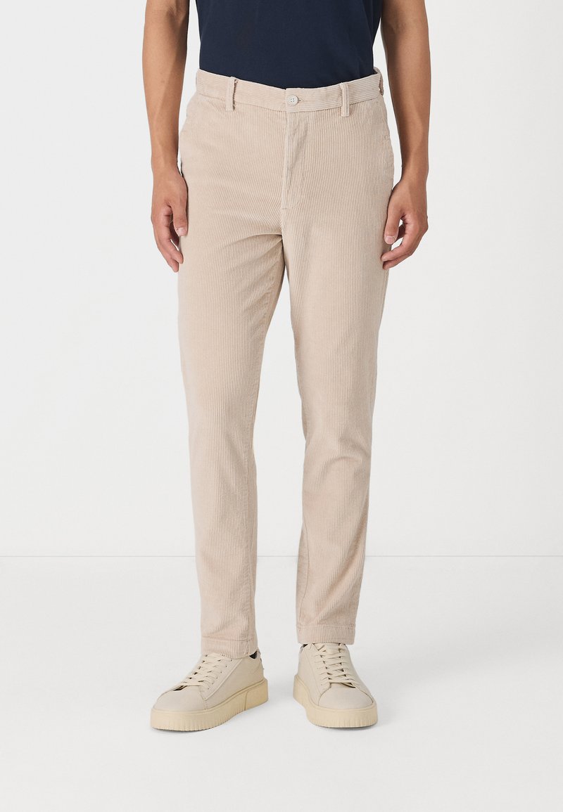 Beige corduroy bukser med lige pasform, der har lodret ribbing, knaplukning og sidelommer. Båret med lysfarvede sneakers.
