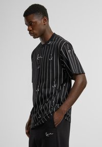 Black t-shirt with vertical white stripes, featuring small white leaf patterns. Short sleeves and a round neckline, made from a smooth fabric.