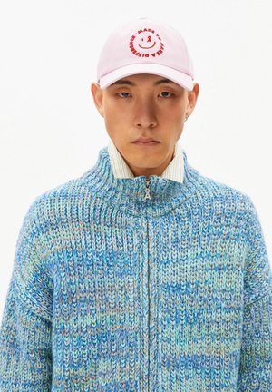 Man met een roze pet met rode tekst en smiley, een blauwgroene gebreide vest met rits en een gestreept overhemd met kraag eronder.