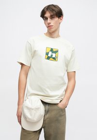 Κρέμα βαμβακερό t-shirt με τετράγωνο λογότυπο σε πράσινο και κίτρινο, συνδυασμένο με ένα ανοιχτόχρωμο καπελάκι που κάθεται στη μέση, φορεμένο με μπεζ παντελόνι.