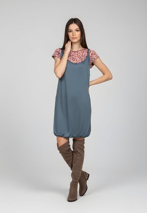 Joven mujer que lleva un vestido azul sin mangas sobre una camisa rosa con estampado y botas marrones hasta la rodilla, de pie con una mano en la cadera.