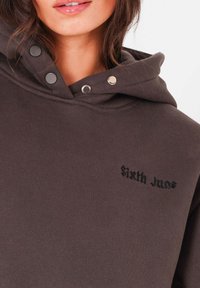 Hoodie marron foncé avec une texture douce, comportant une capuche à cordon, des boutons-pression et le texte brodé "Sixth June" sur la poitrine.
