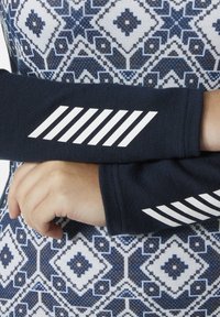 Marinefarbene Armschläuche mit weißen diagonalen Streifen, aus weichem Stoff, kombiniert mit einem gemusterten blau-weißen Hemd mit geometrischen Designs.