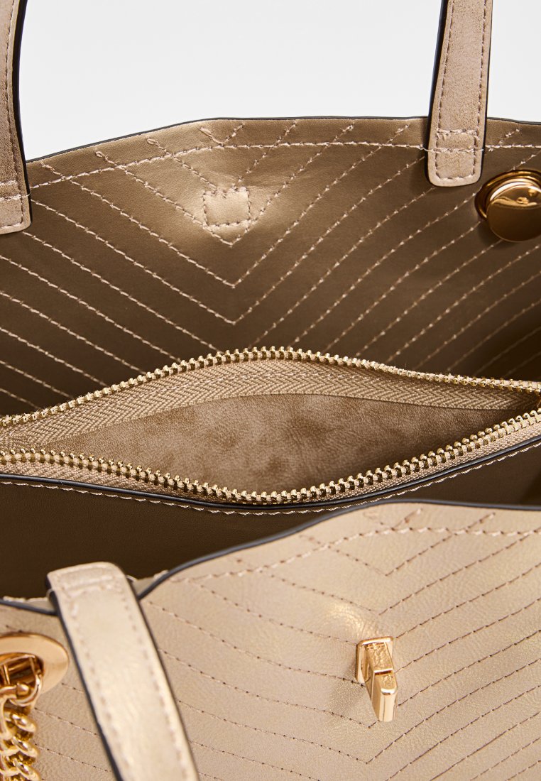 Bolso de mano beige con un patrón en chevron acolchado, herrajes dorados y un bolsillo interior con cremallera forrado con tela suave.