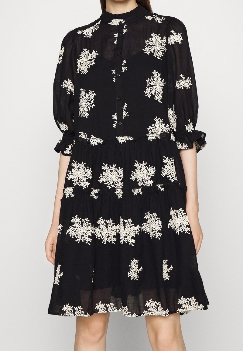 Robe noire longueur genou avec broderie florale blanche, devant boutonné, manches trois-quarts bouffantes et jupe à volants superposés.