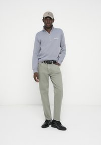 Γκρι φούτερ με φερμουάρ, ανοιχτό πράσινο παντελόνι, μαύρα παπούτσια και μπεζ καπέλο. Απλός σχεδιασμός με ελάχιστο λογότυπο στο φούτερ.