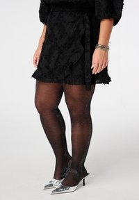 Zwarte kanten rok met een ruchesrand, gecombineerd met zwarte doorschijnende panty’s met zilveren glans en puntige zilveren stilettos.