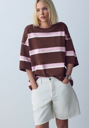 Blonde Frau, die ein übergroßes braunes Hemd mit rosa und weißen Streifen sowie weiße knielange Shorts trägt, die Hände in den Taschen hat und seitlich schaut.