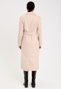 Beige long coat with a belted waist, notched collar, and side split, made from a textured fabric. Worn over black knee-high boots.