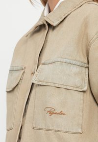 Beige denimjakke med krage, to frontlommer og subtile sømdetaljer med brodert logo.