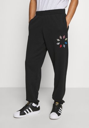 Person trägt schwarze Jogginghose mit bunten Adidas Trefoil-Logos in einem Kreis am Oberschenkel, weißes T-Shirt und schwarz-weiße Adidas-Sneaker.