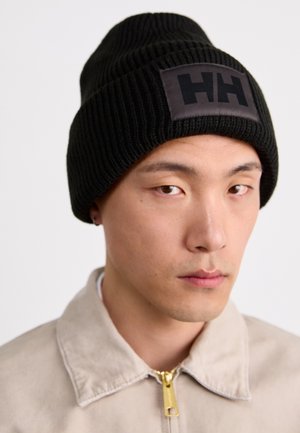 Joven con gorro de punto negro acanalado con el logo "HH" y chaqueta beige con cremallera sobre un fondo blanco liso.