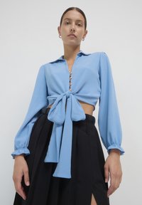 Blusa corta azzurra chiara con fiocco frontale, dettaglio dei bottoni e maniche lunghe; abbinata a una gonna nera a vita alta. Texture liscia.