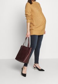 Mujer embarazada vistiendo un suéter mostaza, jeans ajustados oscuros, tacones negros, sosteniendo un gran bolso burdeos, de pie frente a un fondo sencillo.