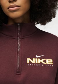 Inimene, kellel on seljas tumepunane Nike neljandiku tõmblukuga dressipluus, millel on rinnal kiri "NIKE ATHLETIC CLUB" ja Nike swoosh logo.