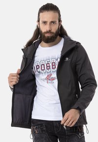 Schwarze Softshelljacke mit Reißverschlusstaschen, Kapuze und Innenfleece-Futter, getragen über einem weißen Graphic-T-Shirt und dunklen Jeans.