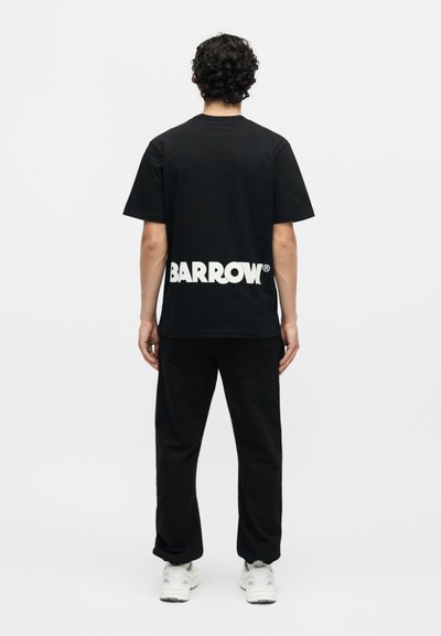 Άνδρας με σγουρά μαλλιά που φοράει μαύρο T-shirt με λευκό κείμενο "BARROW" στο κάτω μέρος της πλάτης, μαύρο παντελόνι και λευκά sneakers, που στέκεται με την πλάτη προς τα εμπρός.