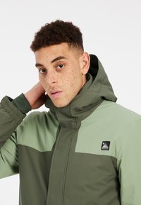 Grün und hellgrüne wasserdichte Jacke mit hohem Kragen, Reißverschluss und Logopatch. Hat eine glatte Textur und verstellbare Bündchen.