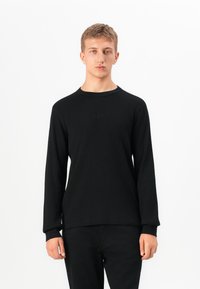Un jeune homme se tenant debout, portant un t-shirt en ribbed noir à manches longues et un pantalon noir, contre un fond blanc uni.
