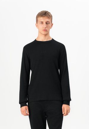Young man standing, wearing a black long-sleeve ribbed shirt and black pants against a plain white background.