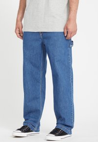 Jeans de mezclilla azul con un corte relajado, que presentan un bolsillo lateral, textura desgastada, y se combinan con zapatillas negras.