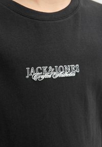 Fekete pamut póló, fehér és ezüst hímzett logóval, amely a mellkason olvasható: "JACK & JONES Crafted Aesthetics".