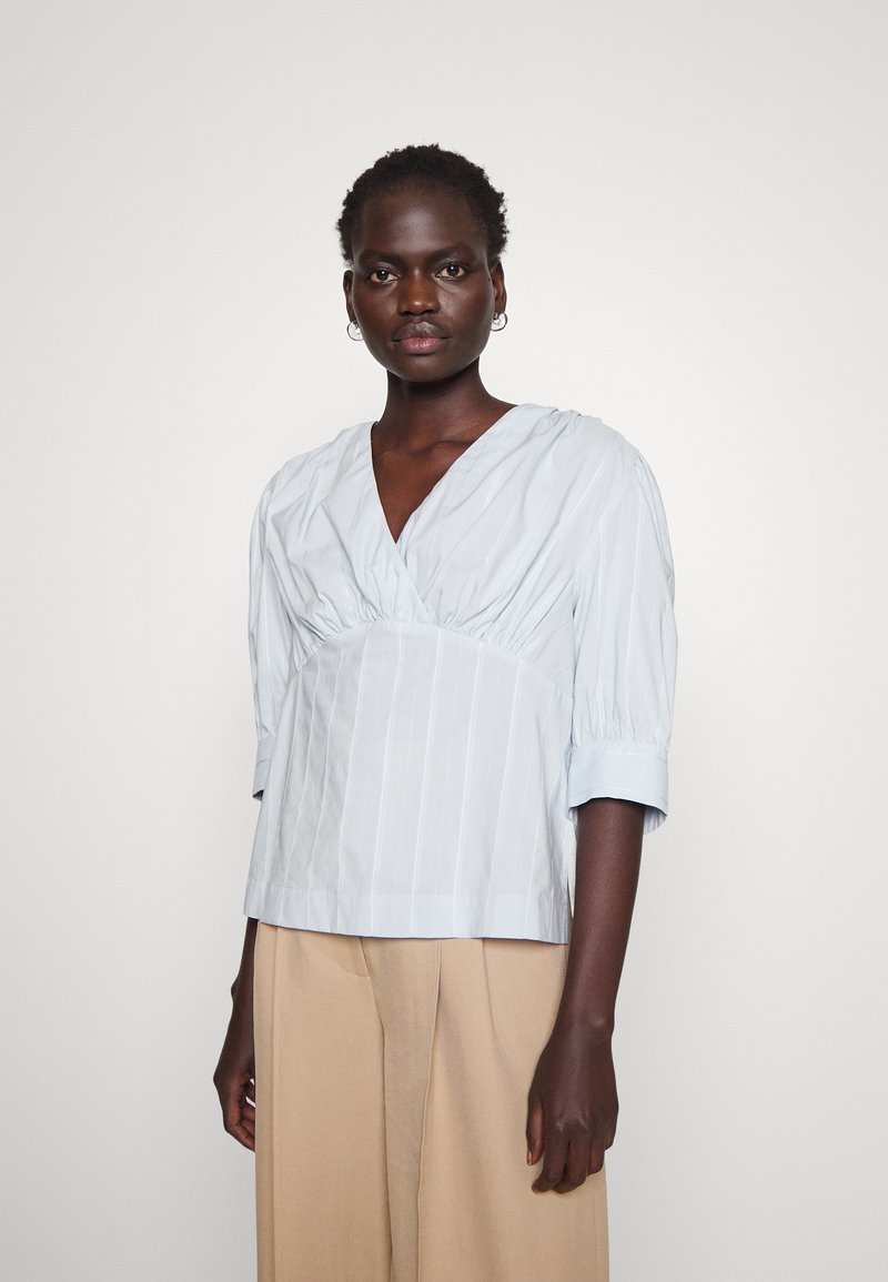 Blusa azul clara em V com busto franzido e mangas 3/4. Com riscas verticais e um corte descontraído, feita de tecido de algodão suave.