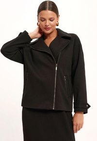 Czarna kurtka zapinana na zamek, wykonana z teksturowanej tkaniny. Posiada duży kołnierz, boczne kieszenie zapinane na zamek oraz mankiety zapinane na guziki. Gładkie wykończenie powierzchni.