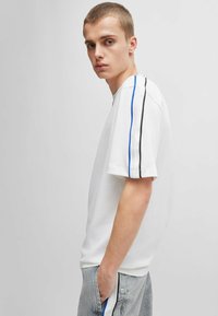 Weißes Baumwoll-T-Shirt mit blauen und schwarzen Streifen an den Schultern. Einfaches Design, kurze Ärmel, lockere Passform, kombiniert mit grauen Jeanshosen.