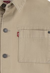 Beige Levi's Hemd aus strukturiertem Stoff, mit Knopfkragen, Brusttasche mit rotem Logosticker und einem schwarzen Druckknopf-Akzent.