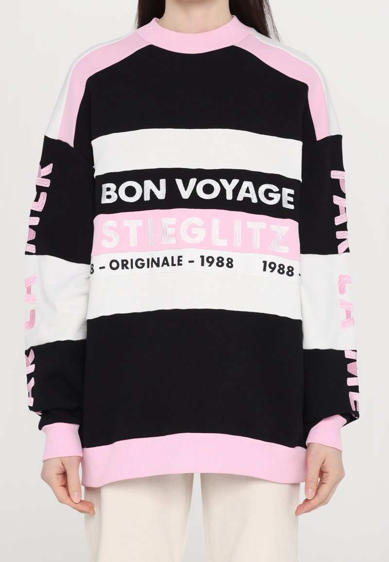 Personne portant un sweat-shirt rayé noir, blanc et rose avec le texte « BON VOYAGE STIEGLITZ ORIGINALE 1988 » sur le devant et les manches.