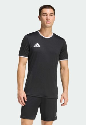 Mann steht und trägt schwarze Adidas-Sportbekleidung mit weißem Saum und Logos auf T-Shirt und Shorts vor einfachem hellem Hintergrund.