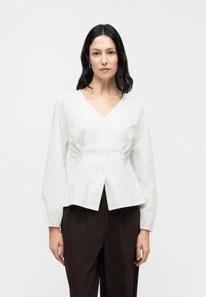 Hvid bluse med lange ærmer og V-udskæring samt en indsnævring ved livet, udstyret med knapper foran og afslappet pasform. Kombineret med mørke bukser.