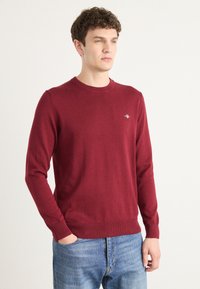 Burgunderfarget strikkegenser med rund hals, lange ermer og ribbestrikkede mansjetter. Liten logo aksent på brystet. Brukt med falmede blå jeans.