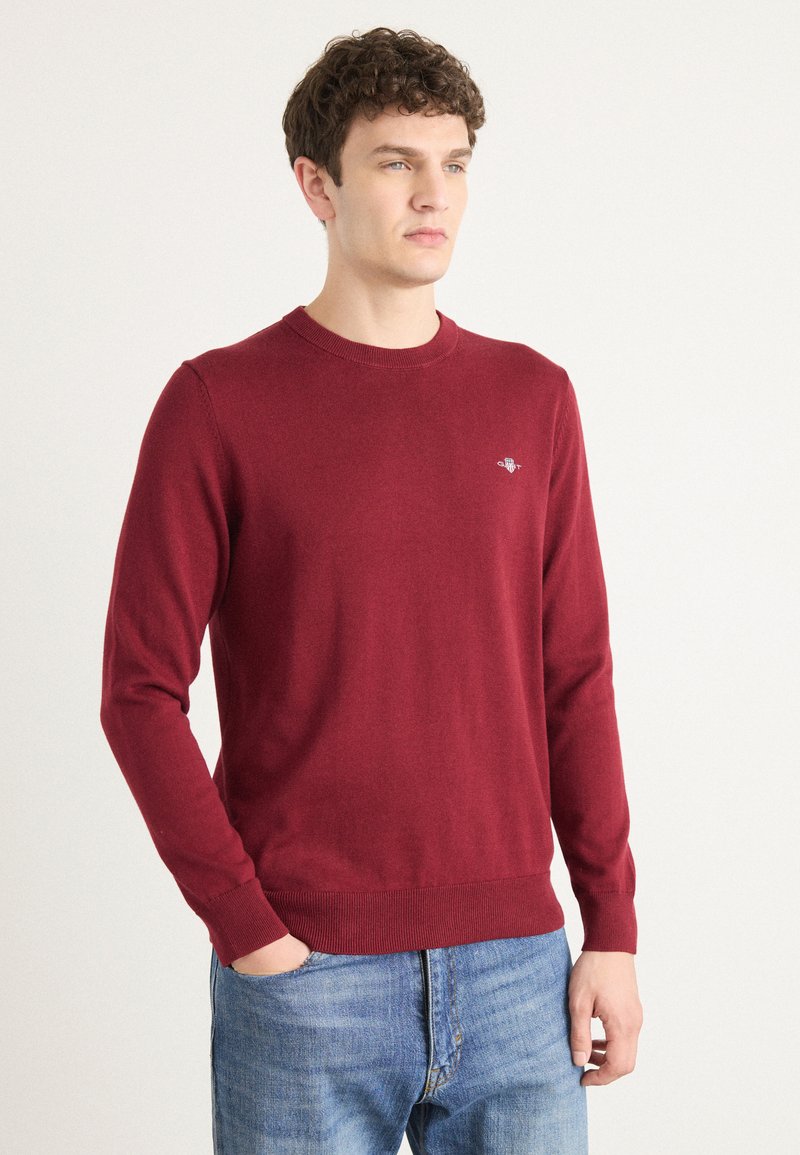 Burgunderfarget strikkegenser med rund hals, lange ermer og ribbestrikkede mansjetter. Liten logo aksent på brystet. Brukt med falmede blå jeans.