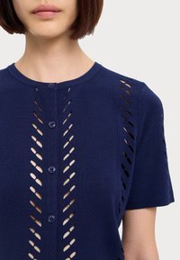 Cardigan tricotat de culoare bleu marin, cu mâneci scurte, având un model orizontal de decupaje și guler circular. Închidere cu nasturi în față.