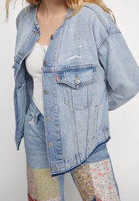 Světle modrá džínová bunda s volným střihem, kovovými knoflíky, dvěma náprsními kapsami a mírnými otvory na látce.
