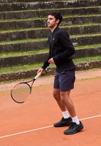 Atleta masculino com uma jaqueta preta de fecho e calções azul-marinho segura uma raquete de ténis num court de argila, usando ténis pretos e meias brancas.