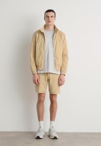 Chaqueta beige claro, camiseta gris y pantalones cortos beige texturizados. El modelo lleva calcetines blancos y zapatillas deportivas grises. Diseño simple y moderno.