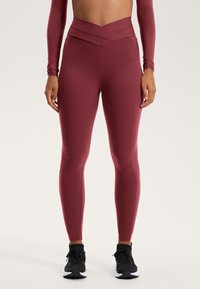 Bordeaux leggings met een hoge, kruis-over tailleband, gemaakt van rekbaar materiaal. Aansluitend ontwerp met stikseldetails, gecombineerd met zwarte sportschoenen.