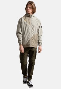 Giacca a vento leggera beige con cappuccio, caratterizzata da dettagli geometrici marroni, chiusura con zip e polsini elasticizzati, abbinata a pantaloni cargo oliva.