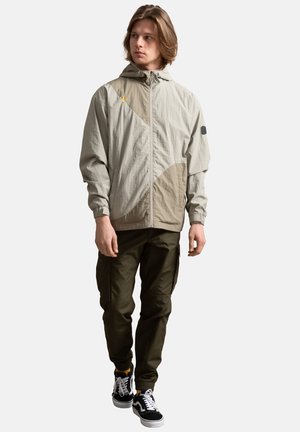 Lichte, beige windbreaker met een capuchon, voorzien van geometrische bruine accenten, ritssluiting en elastische manchetten, gecombineerd met olijfgroene cargobroek.